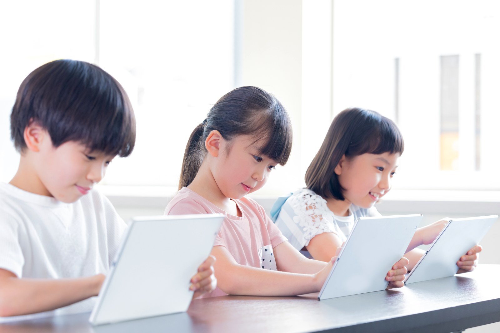 机に並んでタブレットを見ている3人の子ども