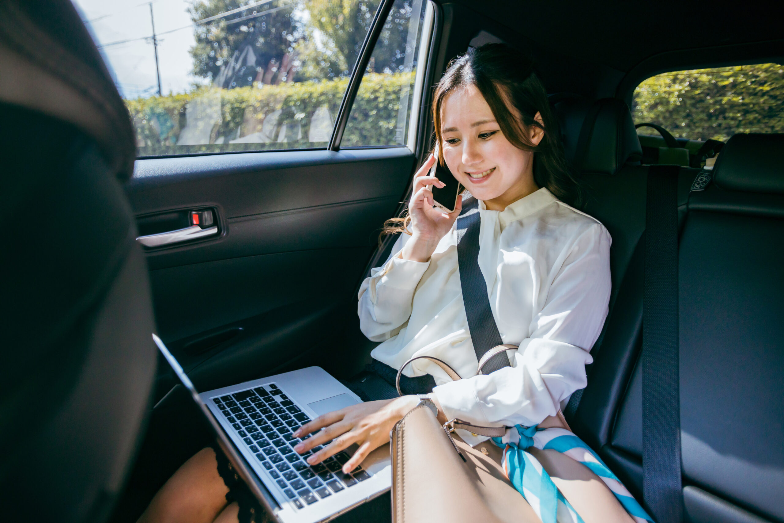 車の後部座席でノートパソコンを開きながらスマホで通話する女性