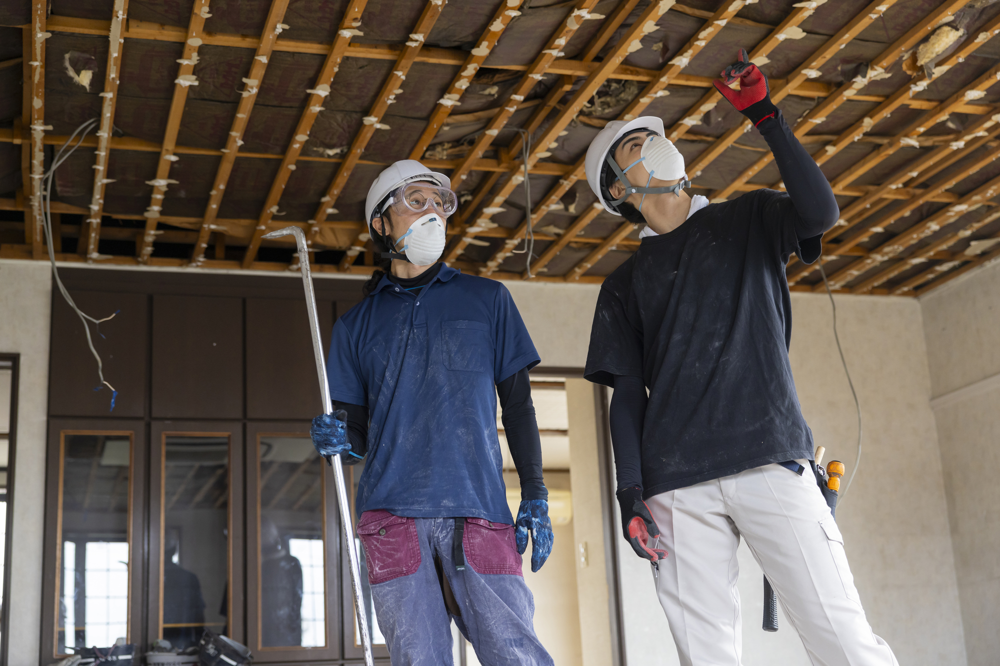天井をはがして解体が進む住宅の室内で、天井を見上げて作業する二人の職人
