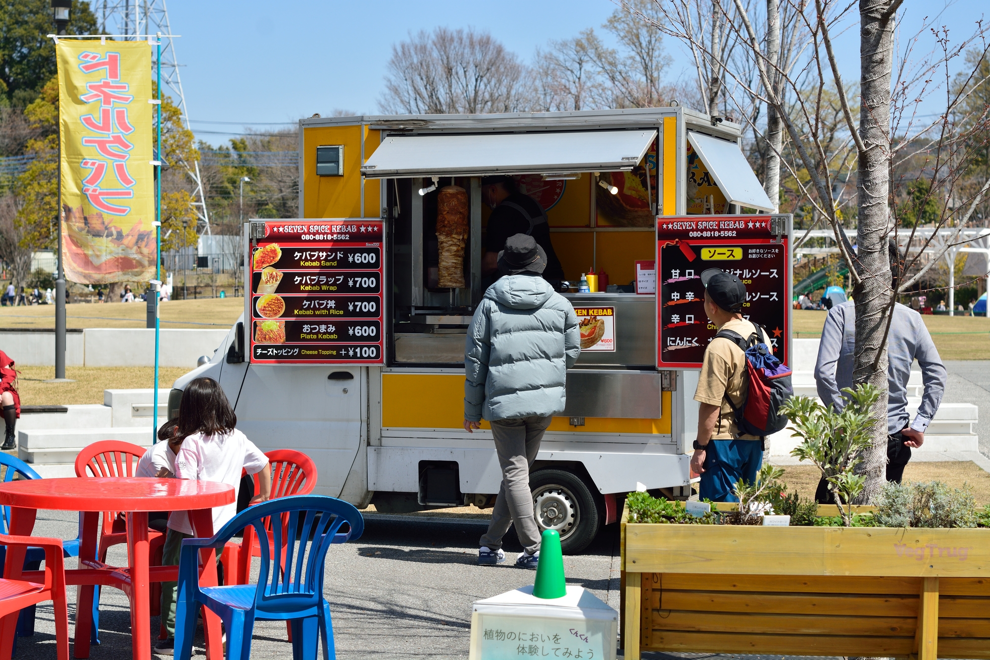 広場に停まった白いキッチンカーに客が並び、手前にはメニュー看板と赤い椅子のイートインスペースがある