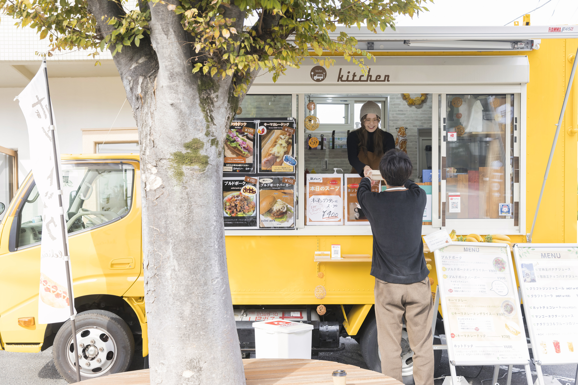 黄色いキッチンカーの窓口で店員が注文を受けており、車の周囲にメニューの看板が並んでいる