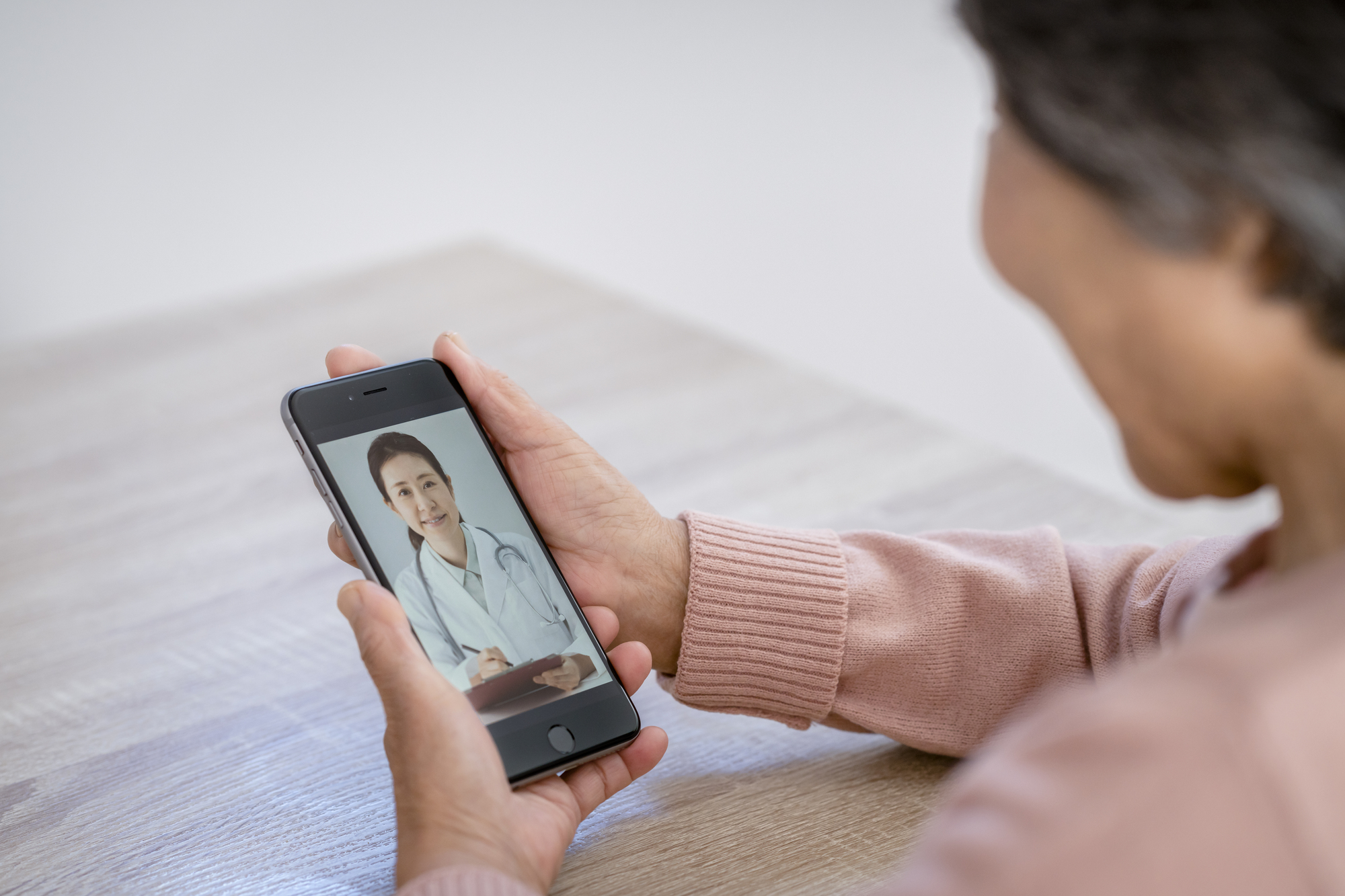 自宅のテーブルでスマートフォンを持ち、画面の中の医師と話す高齢の女性の手元